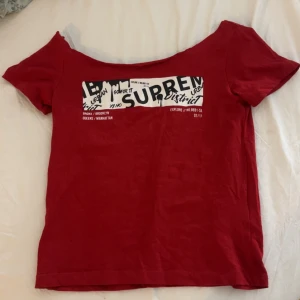 Röd offshoulder topp med tryck - Säljer en röd offshoulder topp från Suprem District med korta ärmar och ett stort vitt och svart tryck framtill. Toppen har en cool urban vibe med text och grafiska detaljer. Perfekt för dig som gillar streetstyle och vill sticka ut.