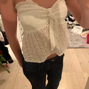 Vit sommartopp - Säljer en söt vit tubtopp med broderade hålmönster och knyt framtill. Toppen är axelbandslös och har en lätt volangkant upptill. Perfekt för varma dagar och ger en fräsch och romantisk vibe.