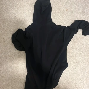 Svart hoodie från MCS med röd logga - Svart hoodie från MCS i storlek S med klassisk huva och snörning. Tröjan har lång ärm och en röd broderad logga på bröstet. Materialet är mjuk bomull och polyester, perfekt för chill dagar. Enkel och stilren design som funkar till jeans eller joggers. Man får med lap, den är äkta