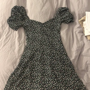 Blommig kortklänning från H&M Divided - Supersöt svart kortklänning med små blommor i vitt, rosa och grönt från H&M Divided. Klänningen har puffiga kortärmar och en smickrande v-ringad hals. Perfekt för dig som gillar romantisk och trendig stil. Materialet är mjukt och lätt, troligtvis bomull eller polyester.