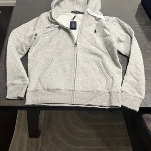 Grå zip Polo Ralph Lauren - Snygg ljusgrå hoodie från Polo Ralph Lauren med dragkedja och huva med vita snören. Tröjan har det klassiska Polo-logot broderat på bröstet och två fickor framtill. Tillverkad i mjuk bomullsmix, perfekt för chill dagar.