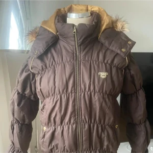 Brun pufferjacka med fuskpäls NY - Brun pufferjacka från JLO med guldfärgade detaljer och dragkedja framtill. Jackan har huva med fuskpäls, quiltad design och mjukt teddyfoder i kragen. 