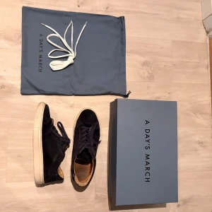 Mörkblå mockasneakers från A Day's March - Snygga mörkblåa sneakers i mocka från A Day's March med rund tå och platt vit sula. Skorna har klassisk snörning och kommer med extra vita skosnören samt originalpåse och låda. Storlek 40 men skorna är stora i storlek så om du har upp mot storlek 42 kan dessa fortfarande passa. Helt okej använda men inget som kommer läggas märke till där ute! Ordinarie pris 1600kr