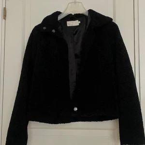 Svart teddyjacka från NLY Trend - Svart teddyjacka från NLY Trend i storlek 32. Jackan har en mjuk och fluffig yta, klassisk krage och långa ärmar. Stängs med knappar framtill och har en croppad passform som ger en cool vibe. Perfekt för dig som gillar stilrena och mysiga plagg.