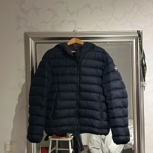 Mörkblå Colmar pufferjacka med huva - Säljer en mörkblå pufferjacka från Colmar med huva och två sidofickor med dragkedja. Jackan har ett stilrent, quiltat mönster och Colmar-logga på ärmen. Perfekt för kalla dagar och har en snygg, modern look.