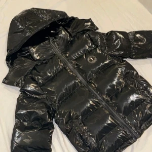 Svart pufferjacka från Trapstar - Säljer en svart, glansig pufferjacka från Trapstar med huva och fet broderad logga på ryggen. Jackan har dragkedja framtill och ett rundat Trapstar-märke på bröstet. Perfekt för dig som vill ha streetstyle med edge och synas i vinter.