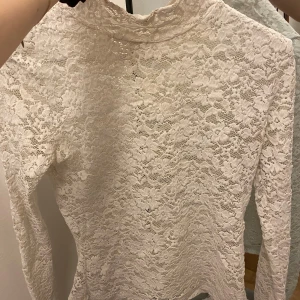 Vit spetsblus med lång ärm - Säljer en vit blus i spets med lång ärm och hög hals. Blusen har ett vackert blommigt spetsmönster och är genombruten för en romantisk look. Perfekt att styla med jeans eller kjol för en trendig outfit.