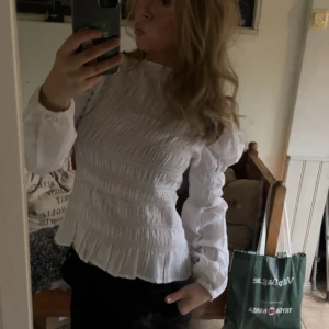 Vit blus med volang och smock - Supersöt vit blus med smock över hela kroppen och volangkant nedtill. Blusen har långärmade puffärmar med resår vid handlederna och en hög hals. Perfekt för dig som gillar romantisk och trendig stil.