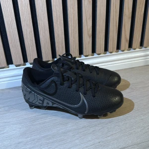 Svarta Nike Mercurial fotbollsskor - Säljer ett par svarta Nike Mercurial fotbollsskor med snygga silverdetaljer och texten 'JUST DO IT' på hälen. Skorna har snörning, platt sula med dobbar och rund tå. Ovandelen är tillverkad i syntetmaterial som ger bra känsla på planen.