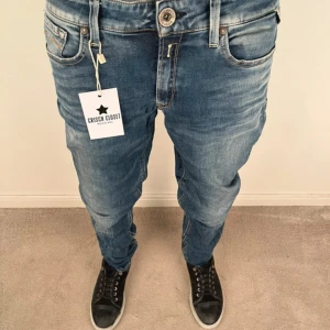 Blå Replay jeans herr, storlek 31/32 - Snygga blå jeans från Replay med klassisk femficksdesign och tydliga slitningar för en cool look. Byxorna har normal passform, raka ben och detaljer som Replay-logga på bakfickan och läderpatch i midjan. Perfekta för dig som gillar streetstyle.
