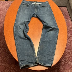 Lee Rider jeans blå W31 L32 - Snygga blå Lee Rider jeans med klassisk femficksdesign och raka ben. Jeansen har mörk tvätt, kontrastsömmar och Lee-logga på bakfickan samt läderpatch i midjan. Tillverkade i slitstarkt denim för en cool och tidlös look. Är mörkare i riktiga livet ljuset får dom se mer grå ut.