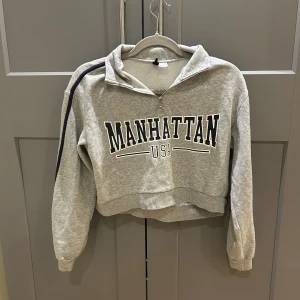 Grå croppad tröja Divided Manhattan - Grå croppad sweatshirt från Divided med texten 'Manhattan USA' framtill. Tröjan har lång ärm med mörkblå rand längs axeln, half zip med ringdetalj och mjuk insida. 