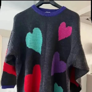  stickad tröja med hjärtan från italienska Gina Benotti - Supermysig mörkgrå stickad tröja från Gina Benotti med stora färgglada hjärtan i rött, lila, turkos och rosa. Tröjan har lila ribbad kant vid hals och nederdel, samt långärmad och loose passform. Perfekt för dig som gillar färg och mönster!