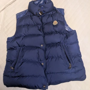 Blå dunväst från Moncler - Snygg blå dunväst från Moncler med hög krage och ikonisk logga på bröstet. Västen har två fickor med dragkedja och quiltad design. Storlek S och i bra skick. 