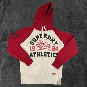 Superdry röd och grå  - Snygg hoodie från Superdry i rött och ljusgrått med stor tryckt logga och text på bröstet. Tröjan har huva med snörning, raglanärmar och en klassisk känguruficka framtill. Perfekt för dig som gillar sportig och avslappnad stil.