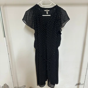 Svart mönstrad klänning från H&M - Svart klänning från H&M med vitt småmönster, v-ringning och korta, luftiga ärmar. Midjan har knytband och tyget är lätt och skönt. Perfekt för dig som gillar en avslappnad men ändå stilren look.