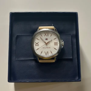 Tommy Hilfiger klocka - Stilren klocka från Tommy Hilfiger med rund silverfärgad boett och vit urtavla med romerska siffror. Klockan har ett guldigt läderband och klassisk design. Levereras i originalask med Hilfiger-logga på urtavlan.