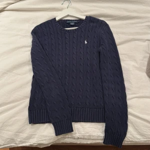 Mörkblå kabelstickad tröja Ralph Lauren - Kabelstickad mörkblå tröja från Ralph Lauren i 100% bomull. Klassisk rund halsringning och diskret broderad logga på bröstet. Långa ärmar och ribbade muddar ger en snygg och tidlös look. Perfekt till jeans eller kjol.