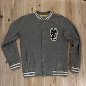 Grå collegejacka från WESC strl.S - Säljer en grå collegejacka från WESC med vita och grå ränder på muddar och krage. Jackan har tryckknappar framtill, två fickor och en snygg broderad logga på bröstet. Perfekt för dig som gillar streetstyle och vill ha en bekväm och stilren look.