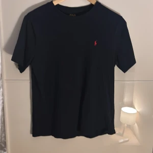 Svart t-shirt från Polo Ralph Lauren - Svart t-shirt från Polo Ralph Lauren med klassisk röd broderad logga på bröstet. Modellen har rund hals och korta ärmar, tillverkad i mjuk bomull som känns skön mot huden. Perfekt för dig som gillar stilrena och tidlösa plagg.