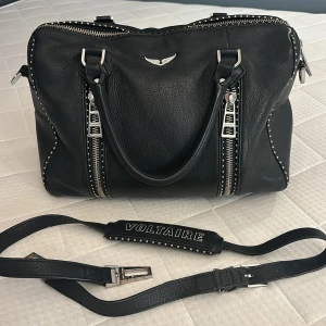 Svart handväska Zadig & Voltaire Sunny BIG - Cool svart handväska i skinn från Zadig & Voltaire med silverfärgade detaljer, dubbla dragkedjor framtill och nitar längs kanterna. Väskan har både handtag och avtagbar axelrem med logga. Perfekt för dig som gillar edgy och stilren design. Fråga så kan jag skicka fler bilder osv. Använt men bra skick! Nypris 6000