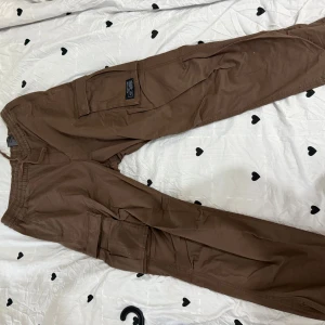 Bruna cargopants med fickor - Säljer ett par bruna cargopants med stora sidofickor och elastisk midja. Byxorna har en avslappnad passform och är perfekta för dig som gillar streetwear och funktionella detaljer. Materialet är mjukt och slitstarkt, vilket gör dem sköna att bära hela dagen.