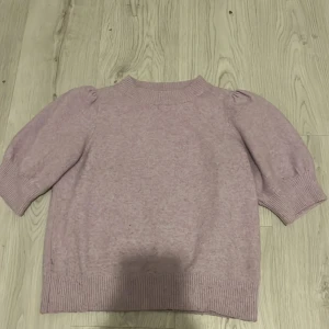 Ljuslila stickad tröja Vero Moda XS - Superfin ljuslila stickad tröja från Vero Moda i storlek XS. Tröjan har puffiga korta ärmar, ribbade muddar och en högre rund hals. Perfekt för lager-på-lager och ger en söt vibe till din outfit. Har en fläck där bak på ryggen som jag tvättar bort innan den skickas
