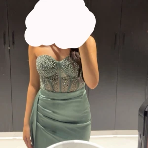 Grön långklänning med pärlor och slits - Säljer en elegant grön långklänning med axelbandslös design och genomskinlig korsett-topp dekorerad med pärlor. Använt bara en gång på ett bröllop. 