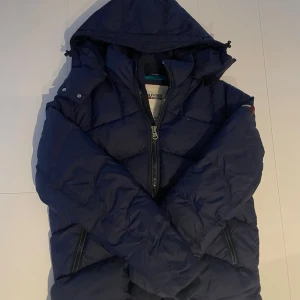 Mörkblå pufferjacka Tommy Hilfiger  - Säljer en mörkblå pufferjacka från Tommy Hilfiger med huva och dragkedja framtill. Jackan har quiltad design, två sidofickor med dragkedja och ett broderat Hilfiger-märke på ärmen. Insidan är ljusblå och jackan har en loose passform. Jackan är använd cirka en gång, ser ut precis som en ny! Storleken är L men jag skulle säga att den är liten för sin storlek, så runt M-S ligger den på
