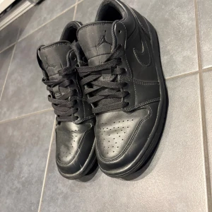 Svarta Nike Jordan 1 sneakers - Säljer ett par helsvarta Nike Jordan 1 sneakers med klassisk siluett och rund tå. Skorna har snörning, perforerad tåbox och är tillverkade i skinn med diskret Jumpman-logga på plösen. Perfekta för dig som gillar stilrena och sportiga sneakers.