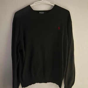 Svart tröja från Polo Ralph Lauren - Klassisk svart tröja från Polo Ralph Lauren med rund halsringning och röd broderad logga på bröstet. Tröjan är långärmad och har ribbade muddar vid ärmslut och nederkant. Perfekt för en stilren och enkel look. Hör av er för frågor eller funderingar!