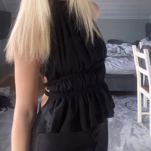 Svart omlott topp med volang - Säljer en svart omlott topp med volangkant och snörning i sidan. Toppen är ärmlös och har en halterneck-design med öppen rygg. Materialet är lätt och lite transparent nedtill, vilket ger en snygg och luftig känsla. Perfekt för dig som gillar trendiga detaljer och en cool siluett. Den är som S och XS