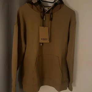 Beige Burberry hoodie med rutigt foder - Säljer en snygg beige hoodie från Burberry med klassiskt rutigt foder i huvan och stora snören. Perfekt för dig som vill ha en stilren och lyxig look med ikoniska Burberry-detaljer.