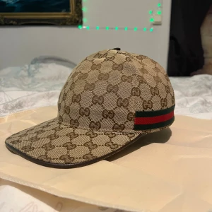 Gucci keps med GG-mönster och ränder - Säljer en beige och brun keps från Gucci med klassiskt GG-monogram över hela kepsen. På sidan finns den ikoniska gröna och röda randen. Justerbar rem baktill i brunt läder. Perfekt accessoar för dig som vill sticka ut med lyxig streetstyle.