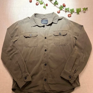 Olivgrön overshirt från Mads Nørgaard - Olivgrön overshirt från Mads Nørgaard med klassisk krage och två bröstfickor med knappstängning. Skjortan har svarta knappar framtill och är tillverkad i mjuk bomull. Perfekt för lager-på-lager och chill stil.