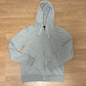 Grå hoodie med dragkedja och huva - Snygg grå hoodie från lager 157 i storlek S. Koftan har hel dragkedja, huva med vita snören och ribbade muddar vid ärmar och nederkant. Hör av vid intresse!