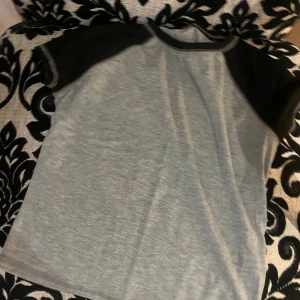 Grå t-shirt med svarta ärmar - Snygg t-shirt i grått med kontrasterande svarta korta ärmar och krage. Enkel och stilren design som passar perfekt till jeans eller shorts. Materialet känns mjukt och bekvämt mot huden, och modellen har en klassisk passform.