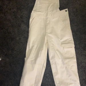Vita hängselbyxor - Snygga vita hängselbyxor med flera praktiska fickor, inklusive benficka med lock. Byxorna har knappstängning i sidan och elastisk rygg för skön passform. Perfekt för dig som gillar funktionella och stilrena byxor. De är aldrig använda. Nypris: 440