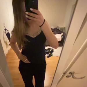 Svart linnetopp med spetsdetaljer - Säljer en svart linnetopp med tunna axelband och snygga beige spetsdetaljer längs halsringningen och sidorna. Toppen har en v-ringad form och är perfekt för dig som gillar en enkel men ändå elegant look. Materialet känns mjukt och följsamt mot huden.