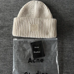Acne studios mössa - Snygg ribbstickad mössa från Acne Studios i en ljusvit ton. Mössan är tillverkad i 100% ull och har ett uppvikt kant med Acne Studios ikoniska ansiktslogga framtill. Perfekt för att hålla dig varm och stilren under kyliga dagar.
