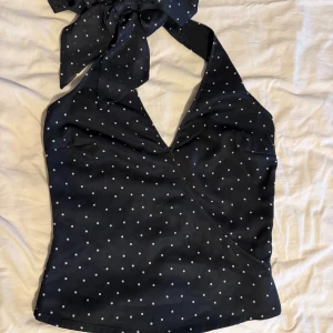 Svart prickig halterneck topp - Svart halterneck topp med vita prickar och omlott-skärning. Perfekt för fest/jul/nyår. Toppen knyts i nacken med breda band och har en v-ringad front. Det finns en dragkedja på sidan. Passar även storlek XS