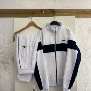 Vit och marinblå vindjacka Lacoste M - Snygg vit vindjacka från Lacoste med marinblåa detaljer och klassisk krokodillogga på bröstet. Jackan har dragkedja framtill, hög krage och elastiska muddar. Perfekt för en sportig och clean look.