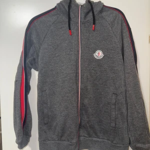 Grå hoodie från Moncler med röd detalj - Säljer en grå hoodie från Moncler med dragkedja och huva. Tröjan har röda och mörkblå ränder längs ärmarna, Moncler-logga på bröstet och snörning i huvan. Materialet är mjuk bomull och polyester, perfekt för chill dagar. Snygg streetwear-vibe!