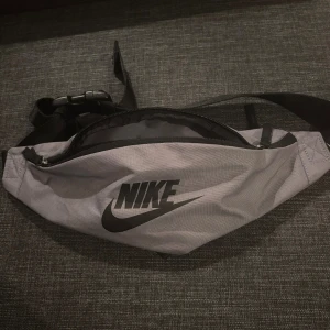 Nike midjeväska - Snygg grå axelväska/midjaväska från Nike med svart logga framtill. Väskan har en enkel, avlång form och justerbart svart band med spänne. 