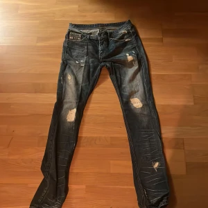 Mörkblå slitna jeans med detaljer - Säljer ett par mörkblå jeans från King Nothing med slitna detaljer och hål på flera ställen. Jeansen har klassisk femficksdesign, coola nitar på myntfickan och kontrastsömmar.