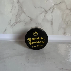W7 Banana Dreams Loose Powder - Banana Dreams Loose Powder från W7 är ett löspuder i en ljusgul nyans som ger en matt finish och jämnar ut hudtonen. Kommer i en rund plastburk med svart lock och gul text. Perfekt för att fixera foundation och ge en fräsch look.