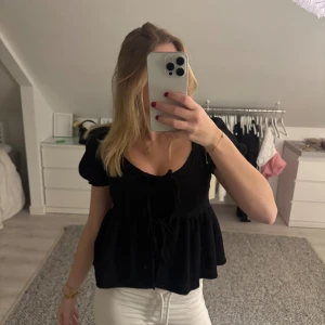 Svart blus med knyt fram - Säljer en svart blus med korta puffärmar och knytning framtill. Blusen har en lös passform och volang nedtill, vilket ger en söt och avslappnad look. Perfekt att matcha med jeans eller kjol.