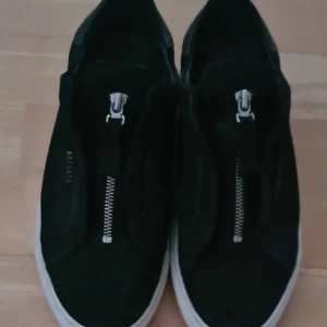 Svarta sneakers från Axel Arigato - Snygga svarta sneakers från Axel Arigato med dragkedja på ovansidan. Skorna har en vit sula och är tillverkade i mocka. Perfekta för en stilren look.