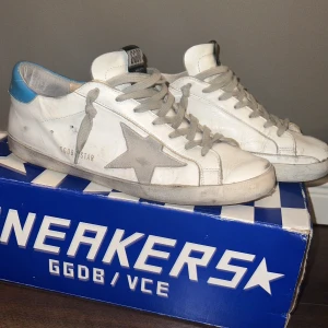 Golden goose - Snygga vita sneakers från GGDB med grå stjärndetalj på sidan och blå häl. Skorna har grå snörning och en klassisk design. Perfekta för en avslappnad stil.