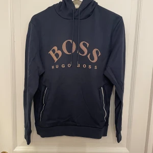 Hugo Boss Hoodie (Gammal Säsong) - Snygg mörkblå hoodie från Hugo Boss med stor logga framtill. Tröjan har en justerbar huva med dragsko och ribbade muddar vid ärmslut och nederkant. Perfekt för en avslappnad stil. Skick 10/10 inga fel på den